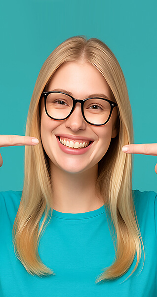 Junge Frau mit Brille lächelt und zeigt mit beiden Zeigefingern auf ihr Gebiss.