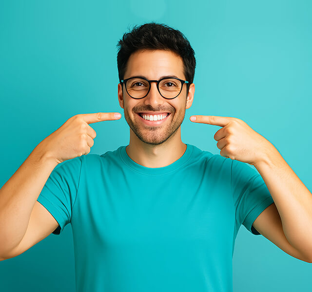 Lächelnde Person mit Brille zeigt mit beiden Händen auf ihre Zähne, türkisfarbener Hintergrund.